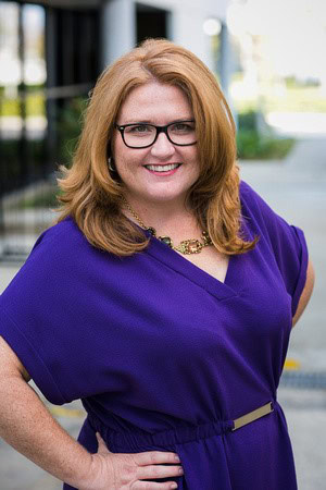 Professional woman smiling outdoors, wearing glasses and a purple dress, representing NAHMA leadersh.