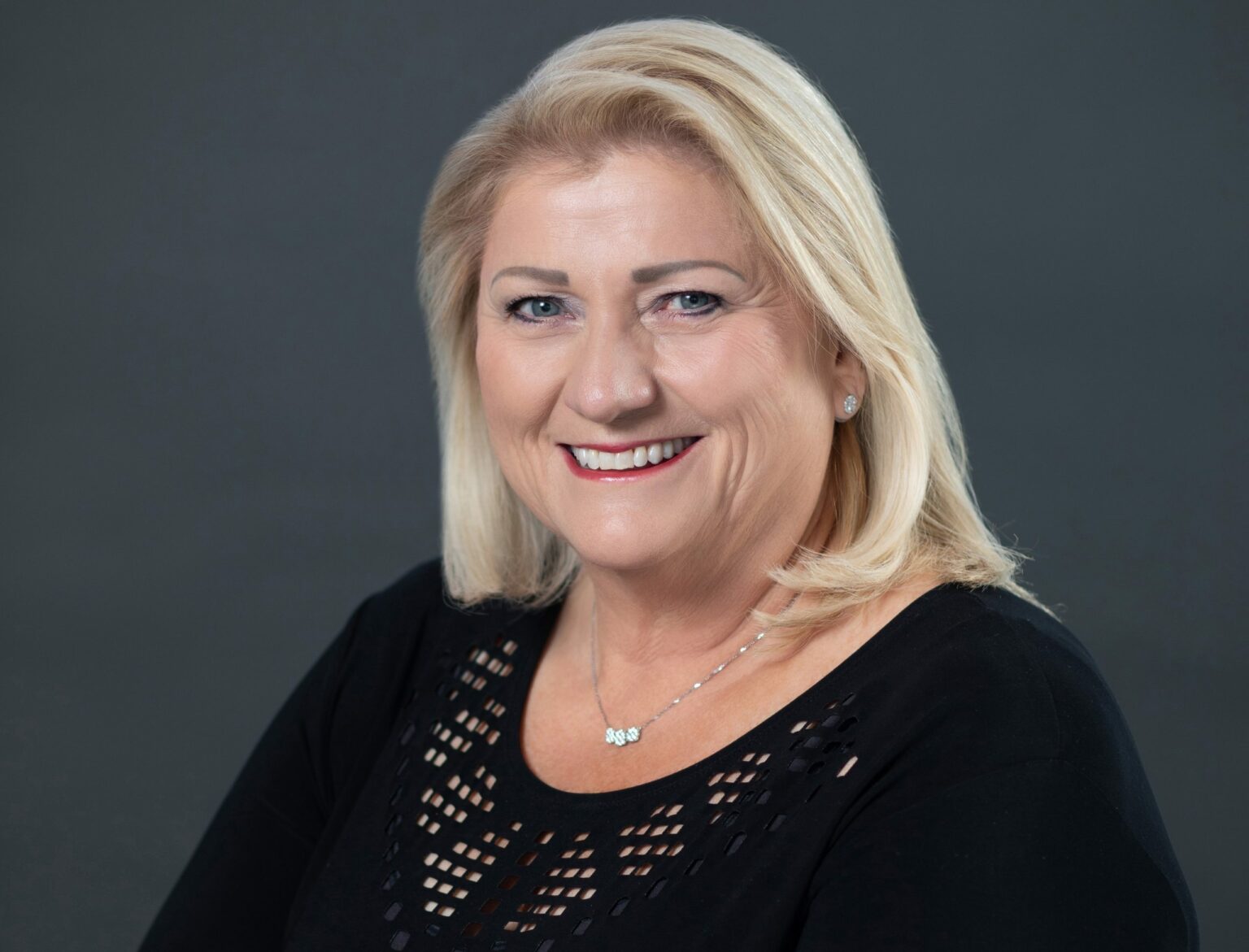 A woman with blonde hair smiling, wearing a black top with lace details, against a gray background.